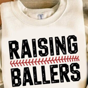 White sweatshirt with 'RAISING BALLERS' text on a wooden surface