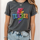 A 3x3 grid showing nine different colorful graphic t-shirts worn by a model. The shirts feature various teacher-themed designs including an apple with the word 'teach', a blue daisy pattern, a rainbow smiley face saying 'BELIEVE SUCCESSION MAKES A DIFFERENCE', a pink heart and book design, a pink and blue 'TEACH A MIDNIGHT' graphic, a rainbow lightning bolt, a patriotic 'TEACHER' design, the word 'teach' in pink script, and a gold glitter-style 'TEACH' graphic.